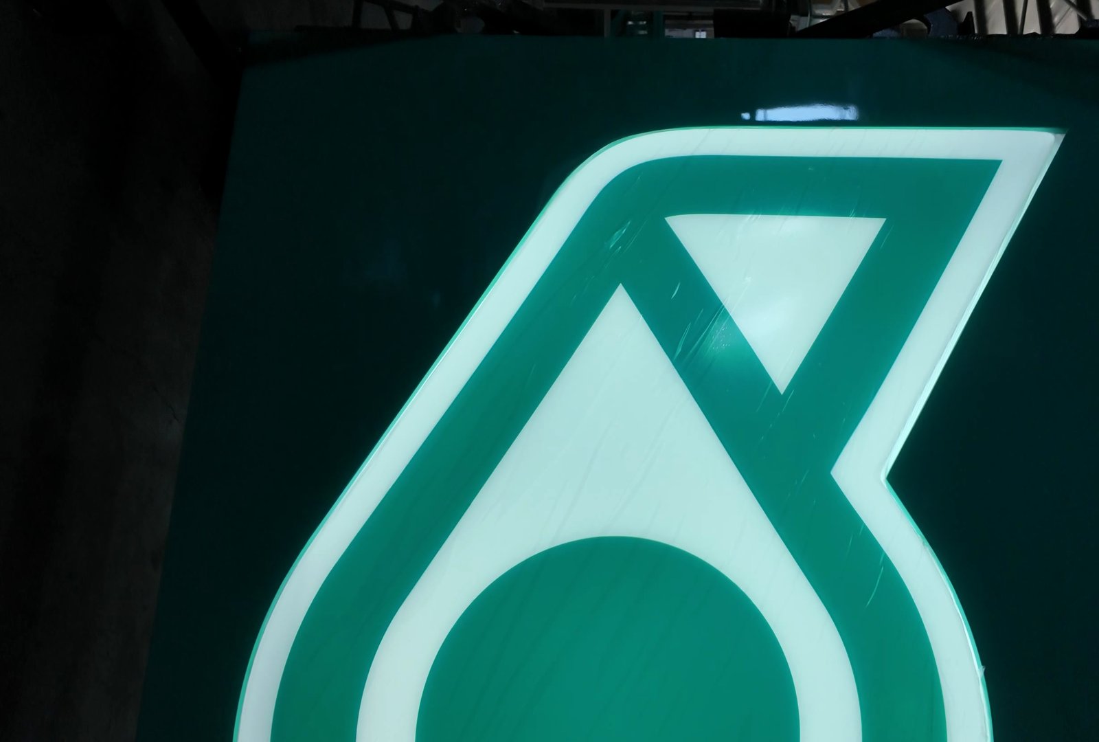 Large petrol-station identity logo panel backlit from within, the white outline and green field both lit evenly across the full panel
